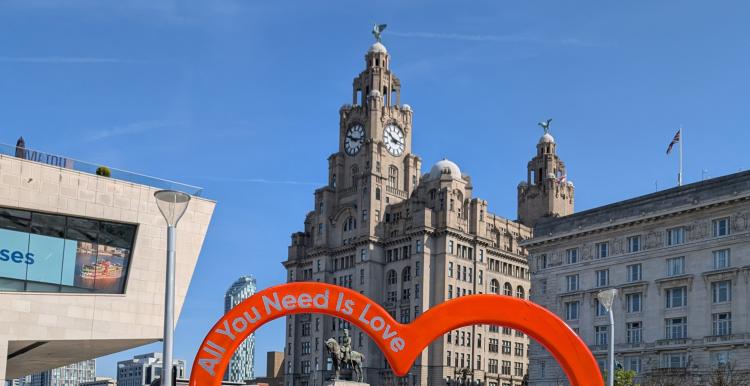 Liver building below a bright blue sky. Red heart in foreground with the words All you need is love.