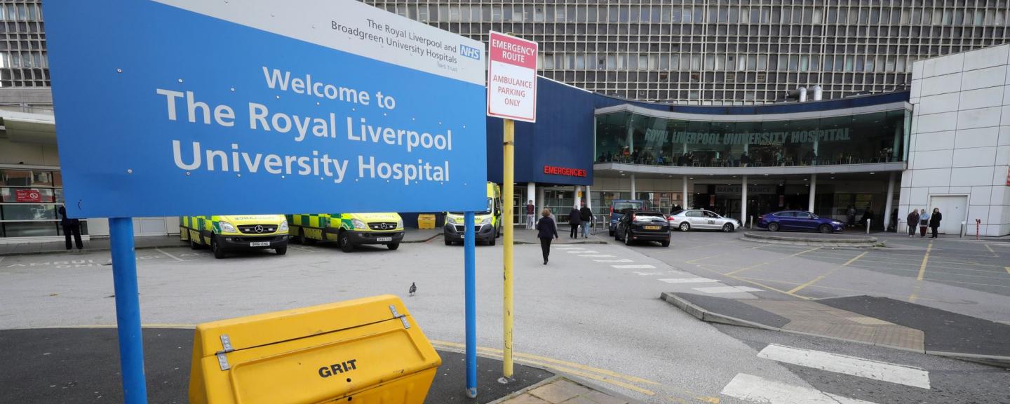 A sign reading 'The Royal Liverpool University Hospital'. In the background is the entrance to the A&E department at the Royal, with ambulances parked outside.