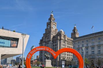 Liver building below a bright blue sky. Red heart in foreground with the words All you need is love.