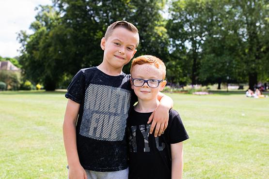 two children standing in the park - the older child has his arm around the younger child's shoulder